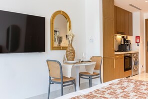 Quarto de hotel moderno com televisão de tela plana, área de refeições com duas cadeiras e uma mesa, cozinha com máquina de café e máquina de lavar roupa.
