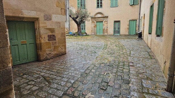 Ein Innenhof mit Kopfsteinpflaster, einem Baum und einem Gebäude mit grünen Fensterläden und einem Ziegeldach.