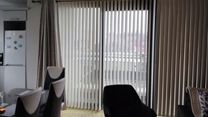 Un salon moderne avec un coin repas, comprenant une grande porte-fenêtre coulissante avec des stores verticaux, un réfrigérateur décoré d’aimants et un canapé noir.