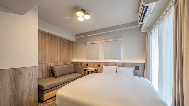 A modern hotel room with a large bed, a built-in bench, a bedside table, and a window with curtains. - Apartment Hotel 11 Dotonbori 8 (Osaka)