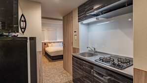 A compact kitchen with a refrigerator, stove, and sink. A bed is visible in the adjacent room. - Apartment Hotel 11 Dotonbori 8 (Osaka)