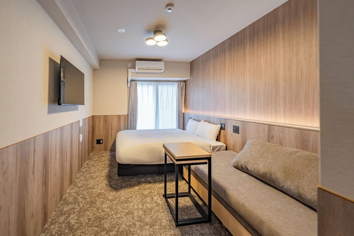 A hotel room with a bed, a sofa, a small table, a TV mounted on the wall, and an air conditioning unit.