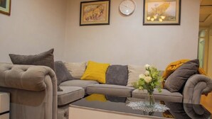 Una sala de estar con un sofá gris, cojines amarillos y grises, una mesa de centro de vidrio con un jarrón de flores, y dos cuadros en la pared.