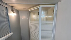 A narrow hallway with a glass-enclosed shower area, a window with blinds, and a light fixture on the wall. - Prime Location Studio with Separate amenities (London)