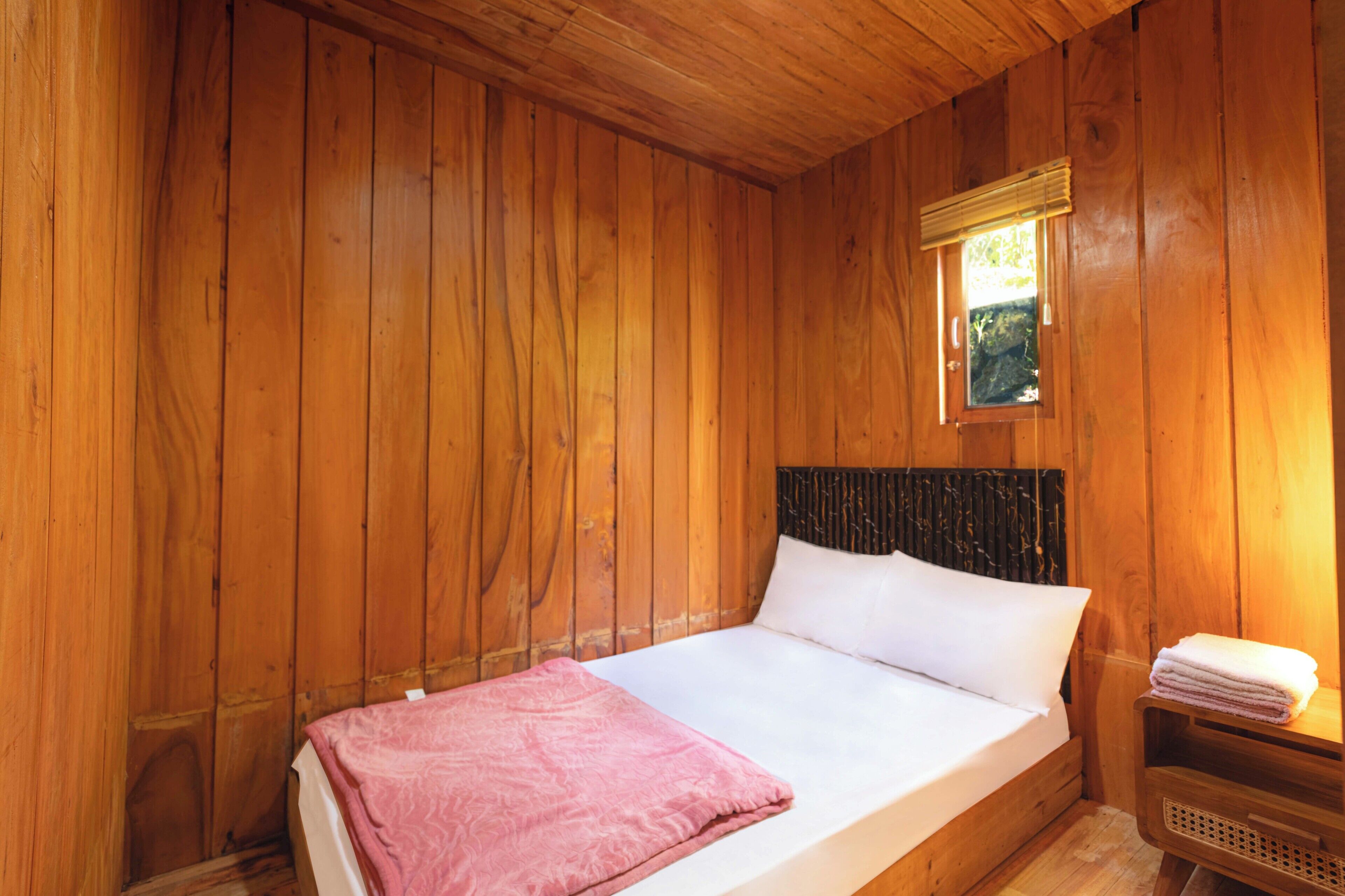 A wooden room with a bed, a window with blinds, and a small table with a towel.