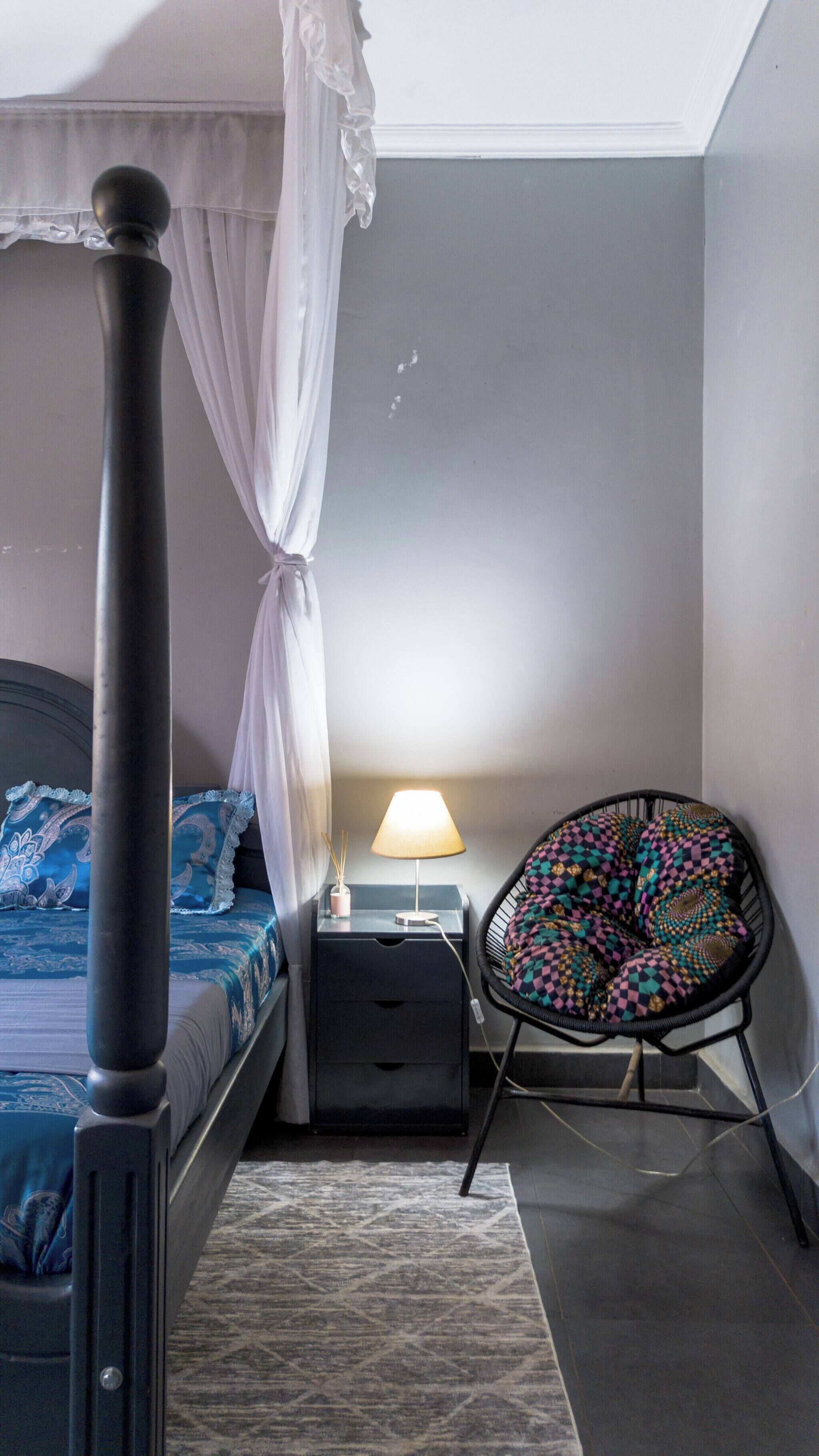 A bedroom with a four-poster bed, a bedside table with a lamp, a chair with a patterned cushion, and a rug on the floor.