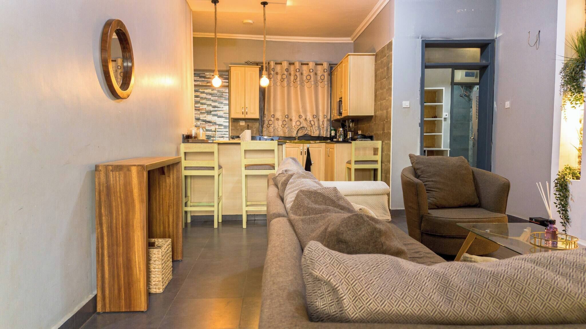 A modern living room with a sofa, a coffee table, and a wooden side table. A kitchen is visible in the background with a dining area.