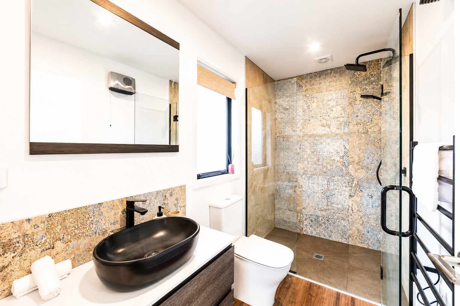 Et moderne badeværelse med et stort spejl, en sort skålhåndvask, et toilet og en bruseafdeling med glasdør.