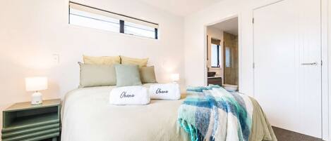 Habitación de hotel con una cama, dos almohadas, una mesita de noche con lámpara, una ventana y una puerta que da a un baño.