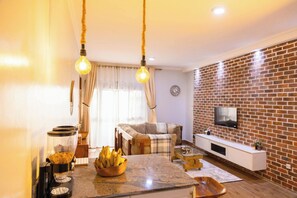 A living room with a brick wall, a sofa, a coffee table, and a TV mounted on the wall. - Charming 2-Bedroom Apartment in Bwebajja, Kampala (Kampala)