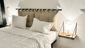 Un dormitorio con cama, almohadas, una mesita de noche con lámpara, un cuadro enmarcado en la pared y una ventana con cortinas.