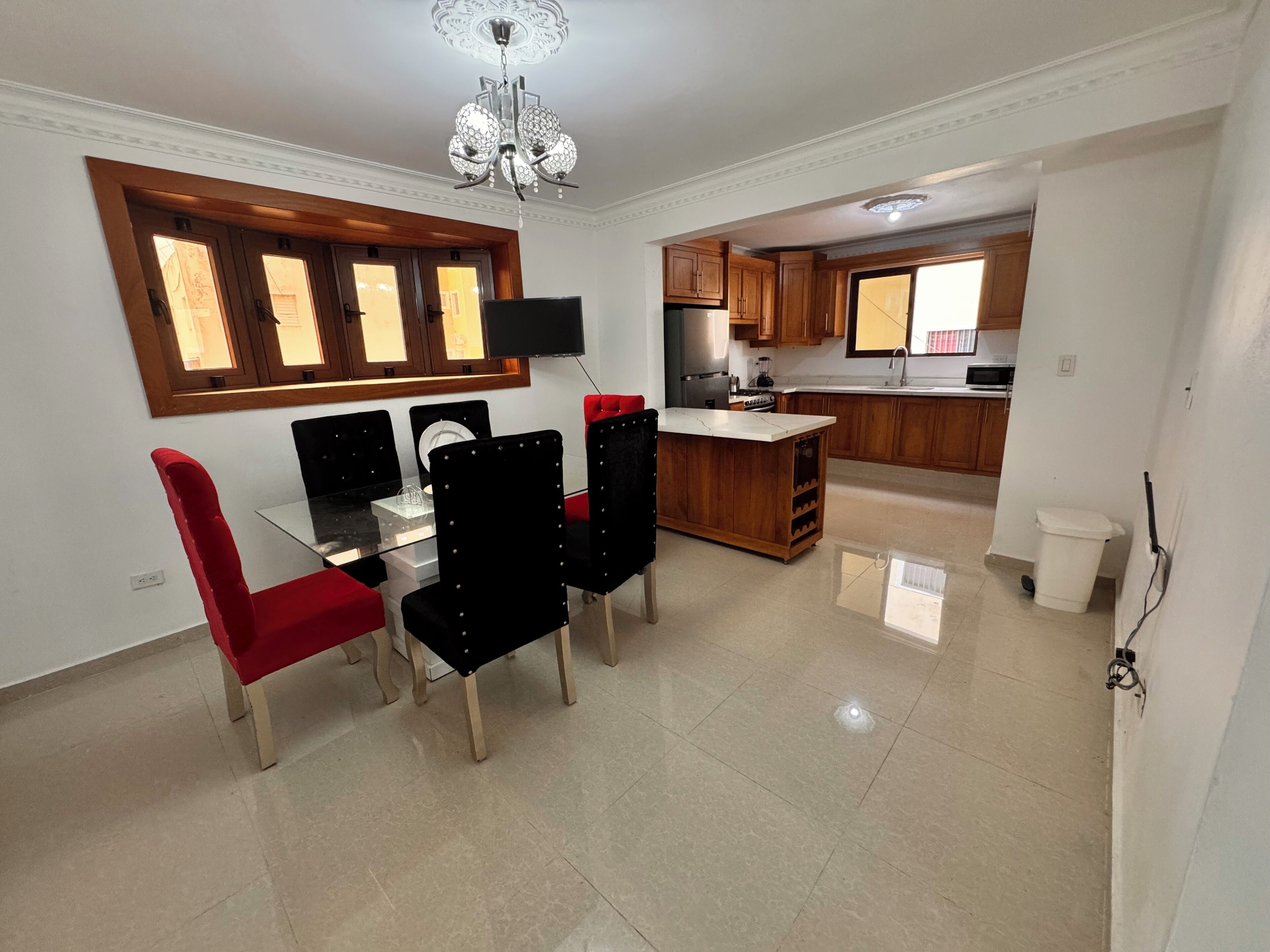 Un comedor con mesa y sillas de vidrio, una cocina con armarios de madera y un televisor montado en la pared.