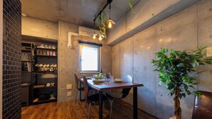 Un comedor con una mesa de madera, sillas, una planta en maceta y una zona de cocina con estantes y utensilios.