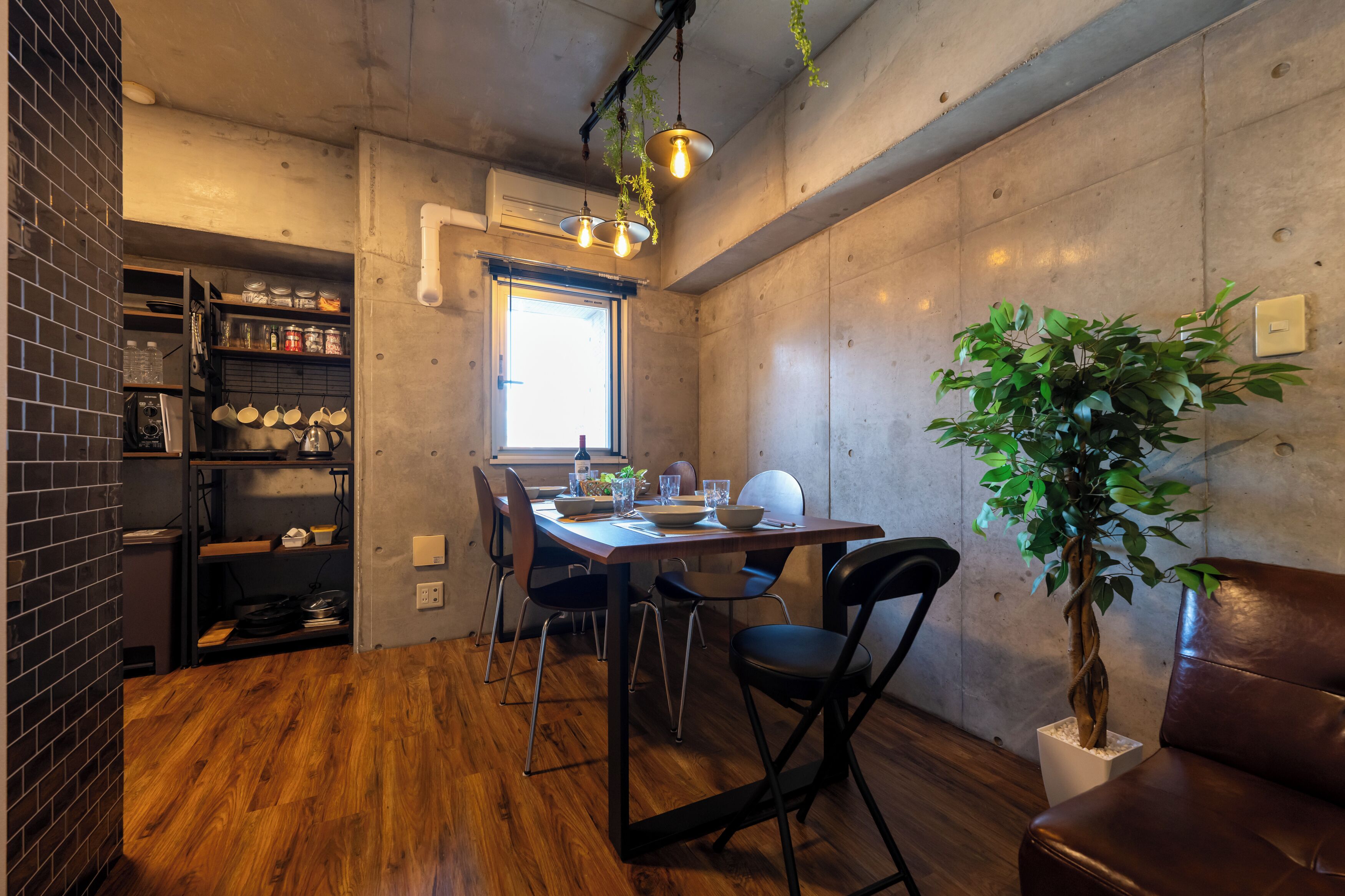 Un coin repas avec une table en bois, des chaises, des luminaires suspendus, une plante en pot et un mur en briques agrémenté d’étagères.