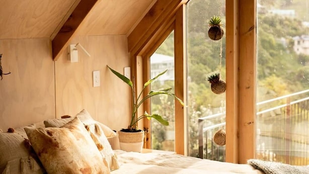 Une chambre confortable avec un plafond en bois, une grande fenêtre donnant sur un paysage verdoyant, un lit recouvert d’un drap blanc et une plante en pot posée sur le rebord de la fenêtre.