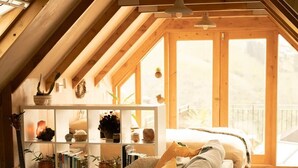 Une chambre à coucher chaleureuse, aménagée en mezzanine, avec un plafond en pente, des poutres en bois, un bureau intégré, un canapé et une grande fenêtre donnant sur l’extérieur.