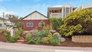 Une maison à deux étages, dotée d’une façade rouge et beige, d’un balcon et d’un jardin bien entretenu où poussent différentes plantes, ainsi qu’un arbuste jaune en fleurs.