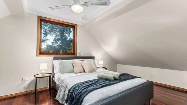 Une chambre avec un plafond en pente, un lit avec une tête de lit grise, deux tables de chevet avec des lampes, une fenêtre donnant sur des arbres et un ventilateur de plafond.