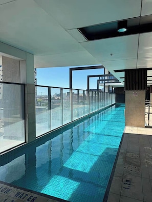 A long indoor swimming pool with clear glass walls, a tiled floor, and a view of the cityscape. - City view focused comfy apartment free parking (Adelaide)