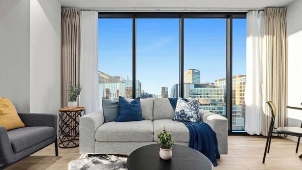 A modern living room with a grey sofa, a black coffee table, and a view of the cityscape through large windows. - City sky view self service apartment free parking (Adelaide)