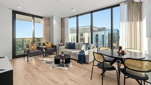 A modern living room with a sofa set, a dining table with chairs, and large windows offering a city view. - City sky view self service apartment free parking (Adelaide)