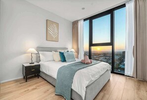 Une chambre moderne avec un grand lit, une table de chevet, une lampe et une vue sur la ville au coucher du soleil.