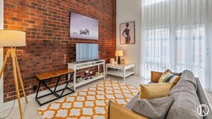 A modern living room with a brick wall, a flat-screen TV, a sofa, a coffee table, and a patterned rug. - Urban Sanctuary Condo in Norwood 2b1b+free parking (Norwood)