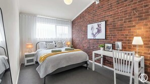 A modern bedroom with a brick accent wall, a bed with white bedding, a desk with a chair, and a hanging light fixture. - Urban Sanctuary Condo in Norwood 2b1b+free parking (Norwood)