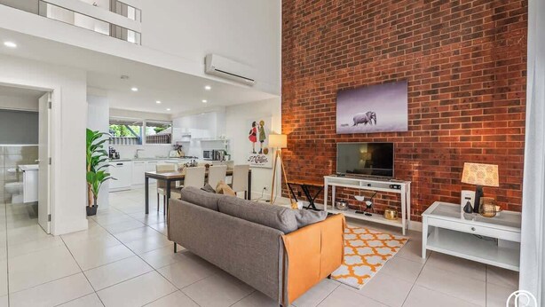 A modern living room with a grey sofa, a television, and a brick accent wall. - Urban Sanctuary Condo in Norwood 2b1b+free parking (Norwood)