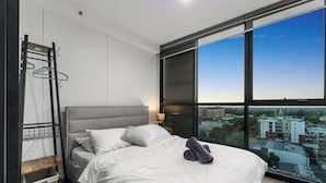 A modern hotel room with a large bed, a view of the city through floor-to-ceiling windows, a built-in wardrobe, and a small shelf with baskets. - Comfort city view apartment Central Adelaide (Adelaide)