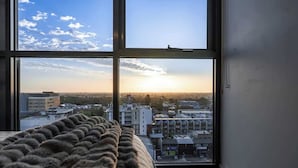 A bedroom with a large window offering a view of a cityscape at sunset. - Comfort city view apartment Central Adelaide (Adelaide)