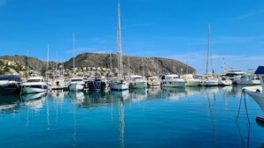 Een jachthaven met diverse boten afgemeerd, een helderblauwe lucht en een heuvelachtig landschap op de achtergrond.
