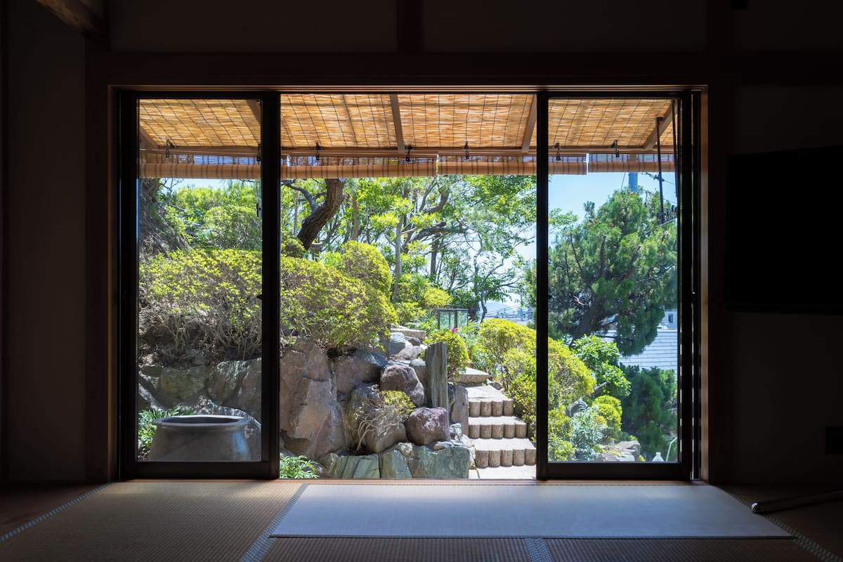 A traditional Japanese room with tatami flooring, a shoji screen door, and a view of a garden with rocks, trees, and a koi pond.
