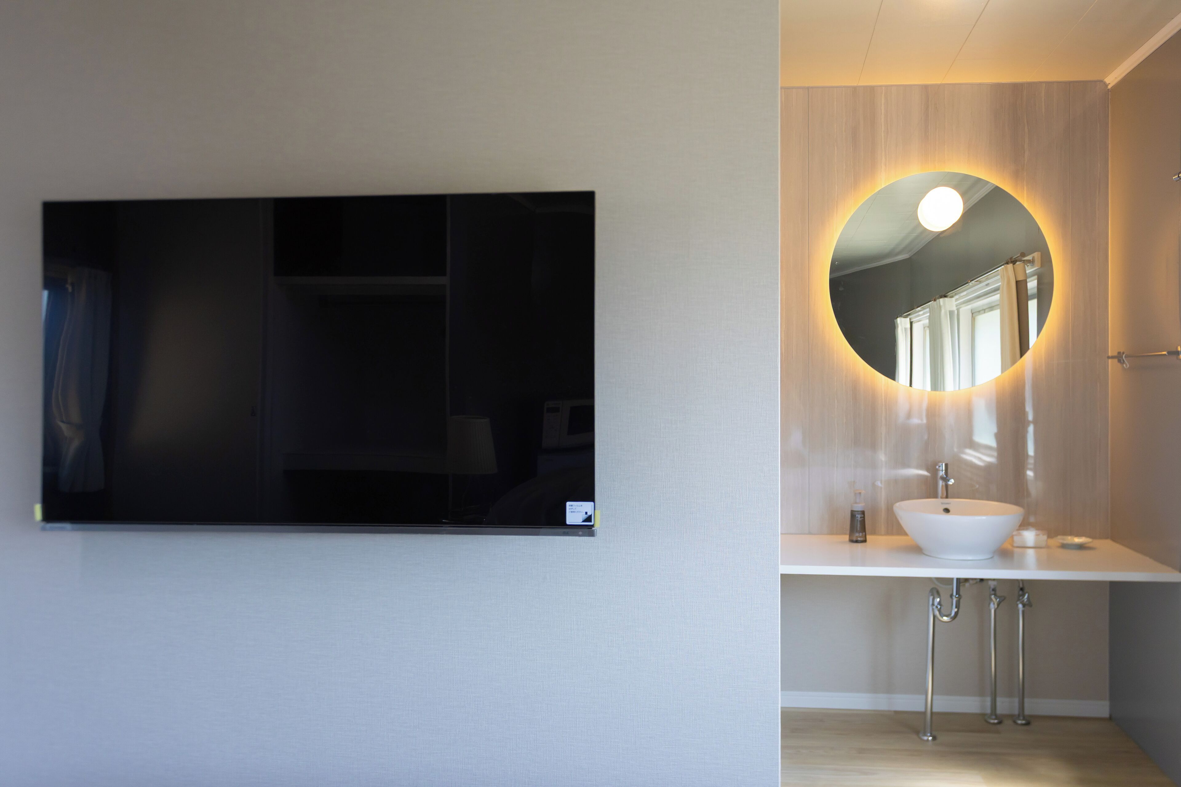 A modern bathroom with a large flat-screen TV mounted on the wall, a round mirror with a light, and a white sink with a vessel basin.