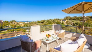 A balcony with wicker furniture, a table with a fruit bowl, and a view of a coastal town. - Villa Aspa in Prines (Rethymno)