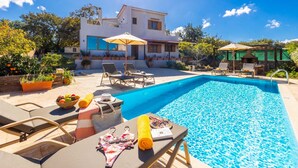 A swimming pool with lounge chairs and umbrellas, surrounded by a garden and a house with a white facade and red tiles. - Villa Aspa in Prines (Rethymno)