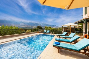 Une piscine entourée de chaises longues et de parasols, le tout délimité par une clôture, avec des montagnes en toile de fond.