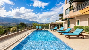 A rectangular swimming pool with blue mosaic tiles, surrounded by a tiled deck. There are several blue lounge chairs with white cushions. The pool is adjacent to a building with a balcony and a view of mountains in the background. - Aloni Villa in Apokoronos (Apokoronas)