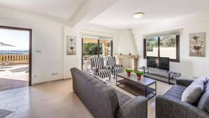 A modern living room with a sofa set, a television, and a fireplace. There is a balcony visible through an open door. - Villa Aqua View in Kissonerga (Pegeia)