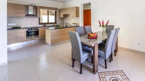A modern kitchen with wooden cabinets, a central island, and a dining area with grey chairs and a table. - Villa Aqua View in Kissonerga (Pegeia)