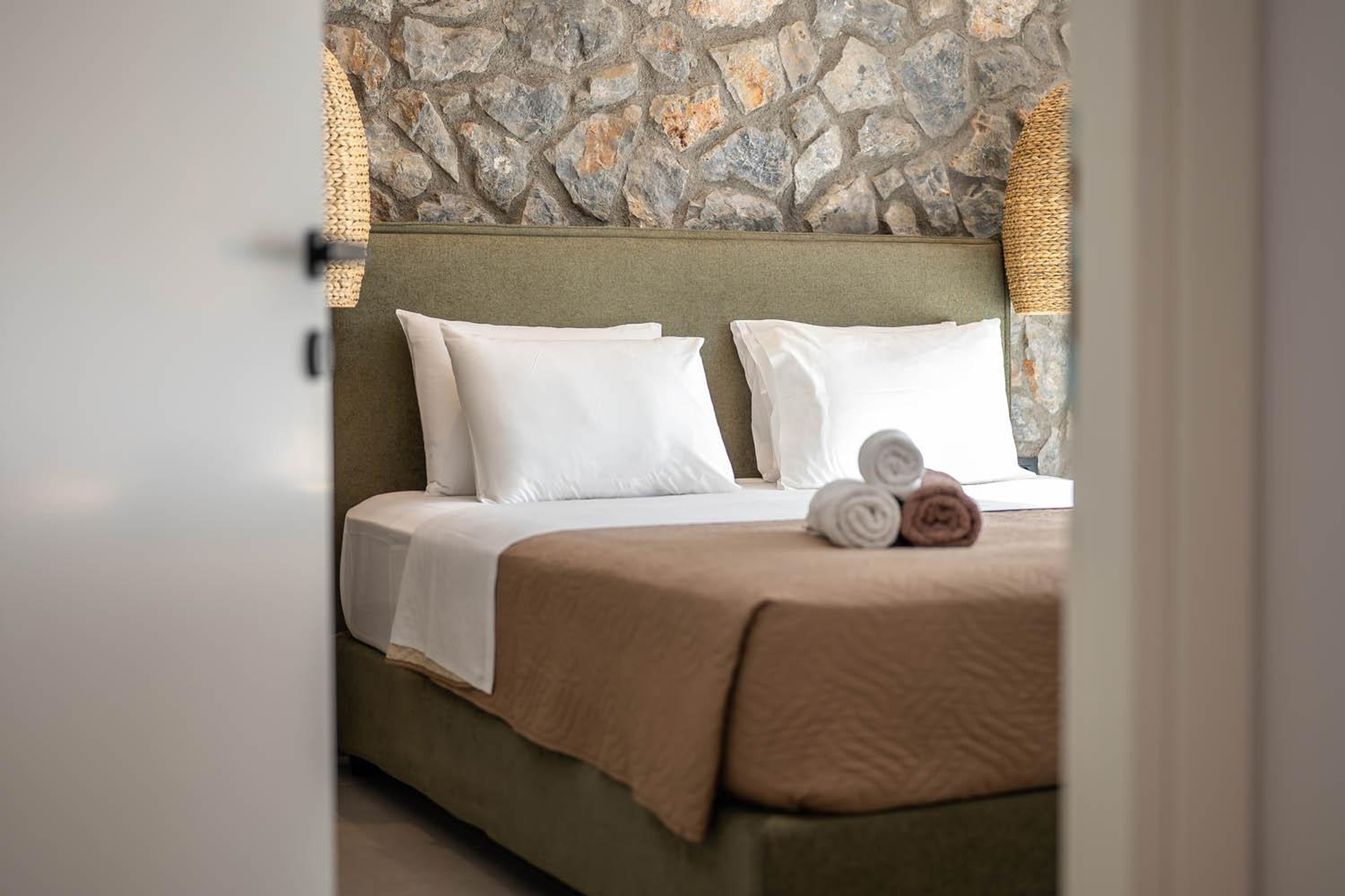 Un lit bien fait, avec du linge de lit blanc et un plaid marron, un mur de pierre en arrière-plan et un panier tressé à côté du lit.