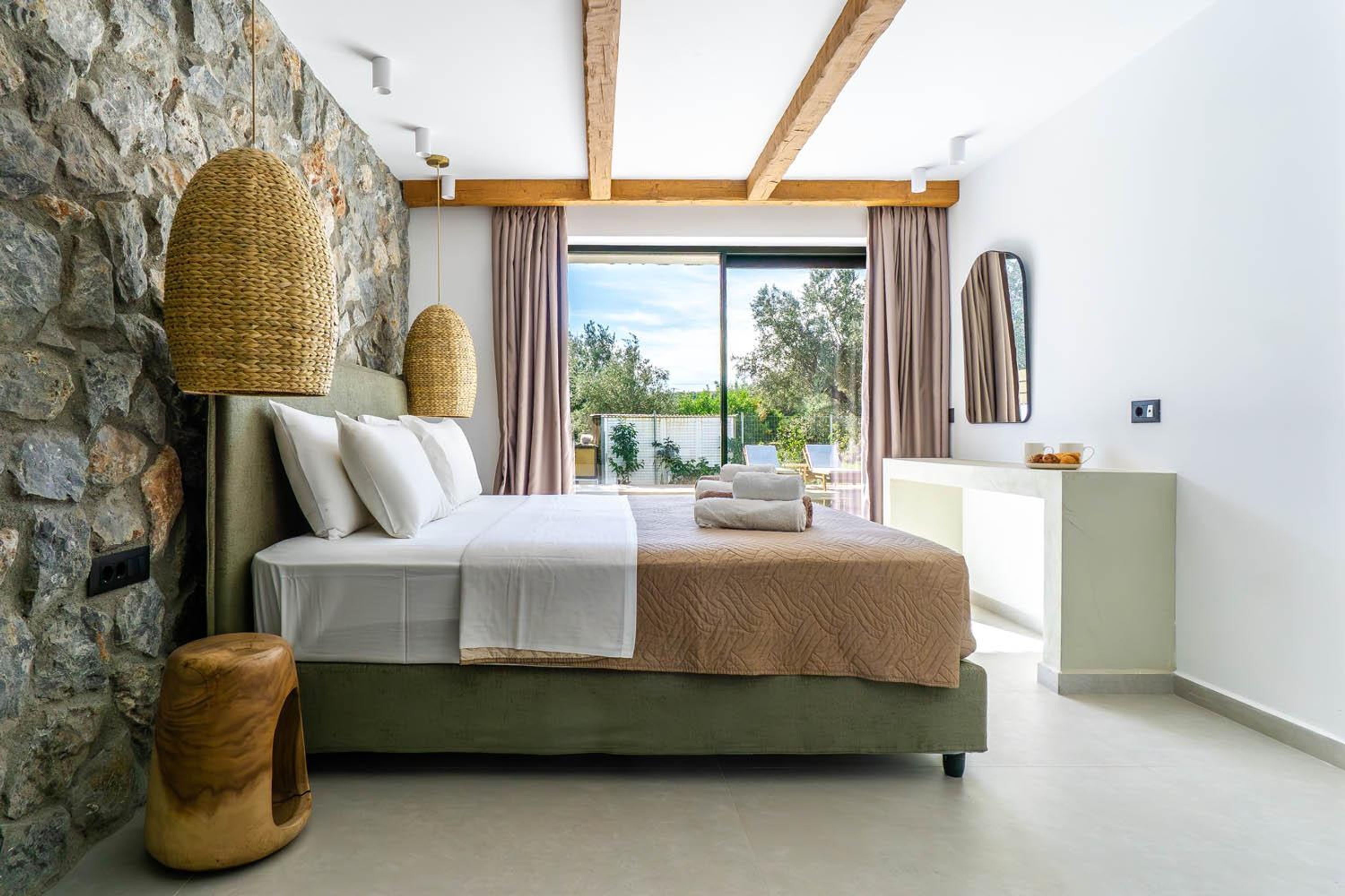 A modern bedroom with a stone wall, a bed with white and beige bedding, a wooden headboard, and a view of the outdoors through large windows.