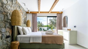 A modern bedroom with a stone wall, a bed with white and beige bedding, a wooden headboard, and a view of the outdoors through large windows. - Villa Aurelia in Afandou (Rhodes)