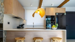 A modern kitchen with wooden cabinets, a central island with stools, and a hanging light fixture. - Villa Aurelia in Afandou (Rhodes)