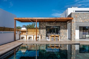 A modern outdoor pool area with a stone wall, wooden pergola, and a barbecue setup. - Villa Amethysta in Afandou (Rhodes)