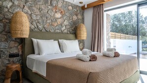 Une chambre avec un mur en pierre, un lit avec une literie blanche et beige, deux lampes en matière tressée et une vue sur l’extérieur.
