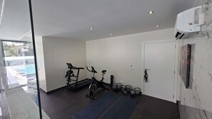 Ein moderner Fitnessraum mit Trainingsgeräten, einer Yogamatte und Blick auf einen Swimmingpool.