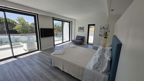 Ein modernes Schlafzimmer mit einem großen Bett, einem Flachbildfernseher, einer Couch und einem Balkon mit Blick ins Grüne.