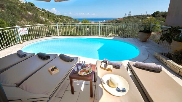 A poolside area with lounge chairs, a table with drinks and snacks, and a view of the sea. - Villa Elpida in Gaios (Paxos)