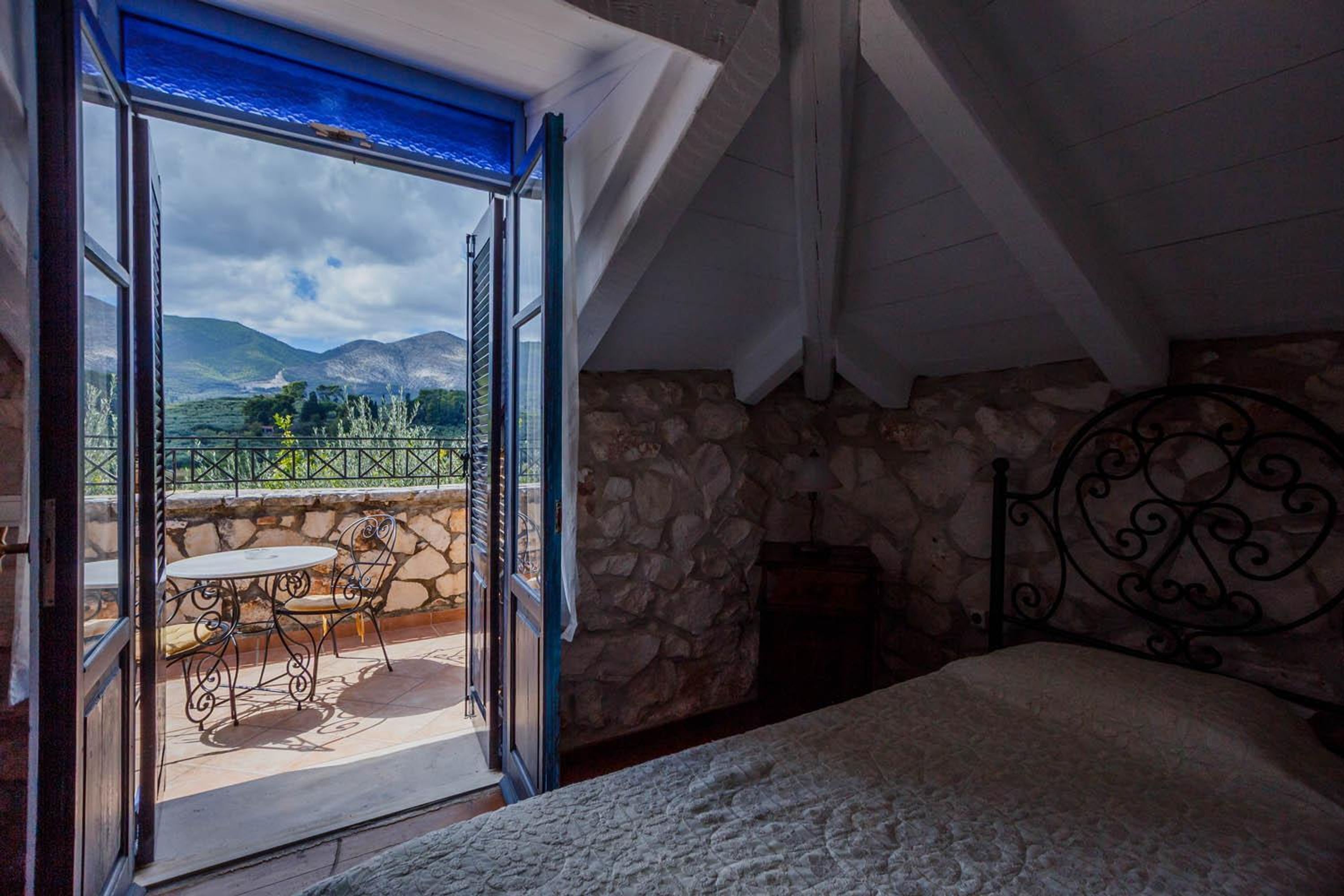 A bedroom with a stone wall, a bed, a balcony with a table and chairs, and a view of mountains.